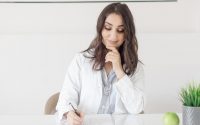 A female nutritionist in a lab coat writing notes with an apple and plant on the desk.