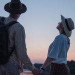 Romantic couple holding hands while watching a sunset at a serene lakeside location, embracing nature.